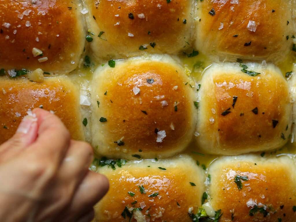 A hand sprinkles sea salt over freshly baked vegan dinner rolls glazed with melted butter and garnished with chopped herbs. The golden brown tops of the rolls are shiny and appetizing.